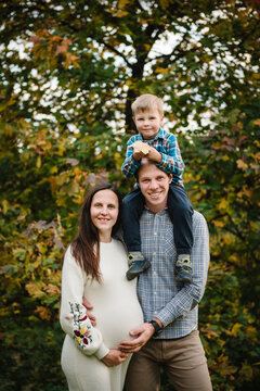 Son sitting on dad's shoulders and hugging mom, happy family portrait in the park in autumn. Pregnant woman and smiling father with child outdoors.