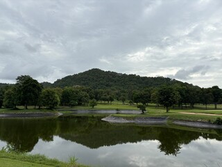 Fototapeta premium Tropical golf course pond with forested mountain reflection under cloudy sky in Thailand