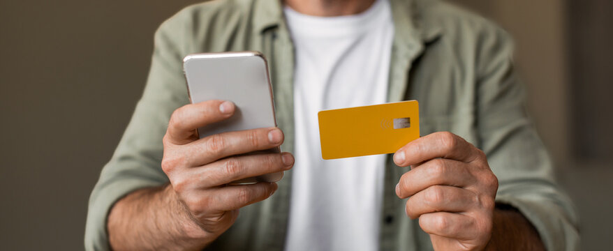 Focused Caucasian Male Adult Holding Smartphone And Yellow Debit Card While Engaging In Online Payment Process At Home Kitchen, Illustrating Modern Financial Transaction Concept.