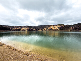 Obraz premium A serene reservoir lake with calm green water, framed by rocky canyons and a pebbled shore under a dramatic cloudy overcast sky.