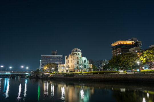 Hiroshima, Japan - 06 March 2020: View of the Atomic Bomb Dome illuminated at night, reflected in the Motoyasu River, with surrounding city buildings and trees under a clear dark sky.