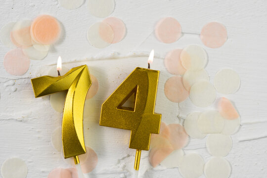 Seventy-fourth anniversary celebration. A golden burning candle in the shape of the number seventy-four on a festive white background with confetti and glitter. Fest
