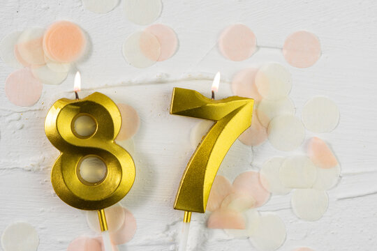 Eighty-seventh anniversary celebration. A golden burning candle in the shape of the number eighty-seven on a festive white background with confetti and glitter.