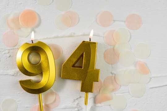 Ninety-fourth anniversary celebration. A golden burning candle in the shape of the number ninety-four on a festive white background with confetti and glitter.
