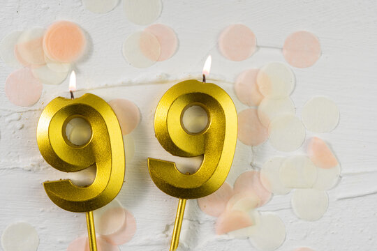 Ninety-ninth anniversary celebration. A golden burning candle in the shape of the number ninety-nine on a festive white background with confetti and glitter.