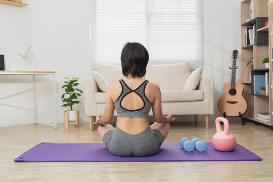 Woman practicing yoga in living room, meditating, sitting cross-legged on purple mat with fitness equipment and guitar, cozy home environment