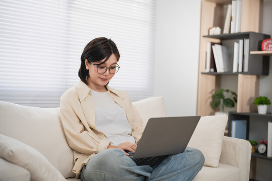 Woman Using Laptop on Couch in Living Room with Bookshelf and Plants in Bright Natural Light