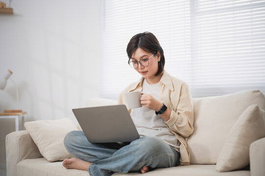 Woman Relaxing on Couch with Laptop and Drinking Coffee in Bright Living Room