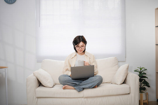 Young woman with glasses sitting cross-legged on sofa using laptop and smartphone in modern living room during daytime