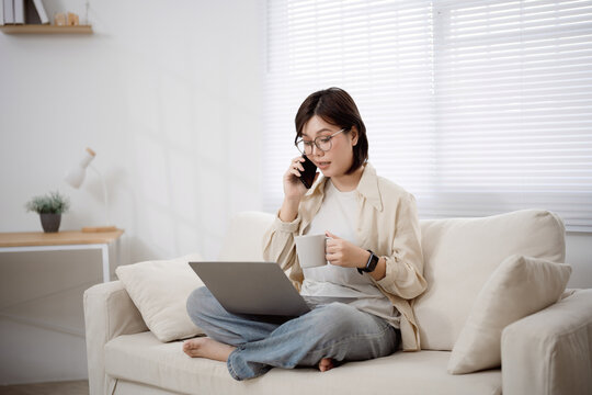 Young woman sitting cross-legged on sofa using laptop computer and talking on cellphone while holding mug in bright modern living room