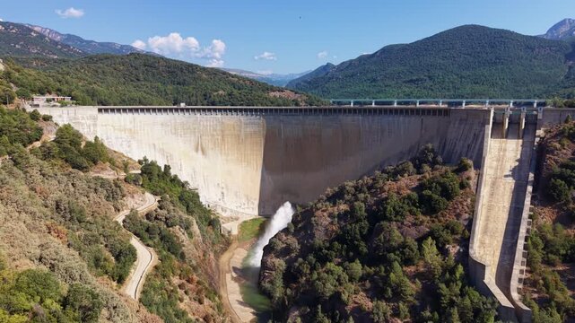 Baells Dam in Catalonia, Spain, releasing water discharge from its base, surrounded by green forested mountains and clear blue sky