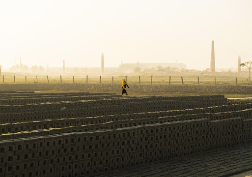 Chattogram, Chittagong Division, Bangladesh - 02 January 2026: View of a woman in a yellow headscarf walking through a brick field with rows of drying bricks and chimneys in the hazy background.