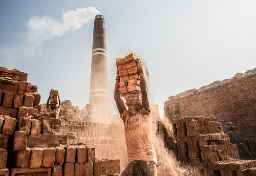 Chattogram, Bangladesh - 02 January 2026: View of a worker carrying a stack of bricks on his head at a brick factory, with a tall chimney emitting smoke in the dusty background.