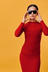 Fototapeta premium Woman in red dress wearing large sunglasses standing against orange background with confident expression and fashion style, studio portrait of female model posing.