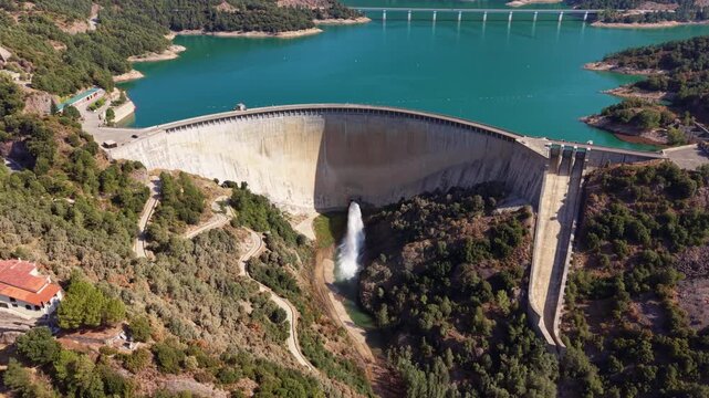 Aerial view of Baells Dam in Catalonia, Spain, showing a powerful water discharge from the concrete arch dam with a turquoise reservoir and forested hillsides
