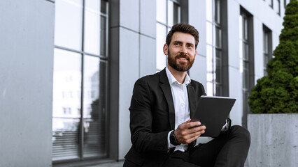 Fototapeta premium Smiling Young Caucasian Man Sitting On Steps Relaxing While Using Tablet In Modern Office Setting, Engaged With Device And Enjoying Break, Capturing Professional Business Concept.