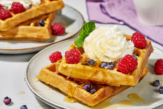 stacked golden brown waffles topped with fresh raspberries and blueberries, whipped cream, and syrup, inviting breakfast dessert option on plates from above close-up.
