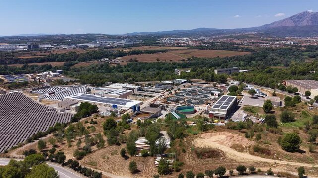 Aerial view of a large drinking water treatment plant with circular clarifier tanks, solar panels on rooftops, surrounded by green landscape and mountains in background