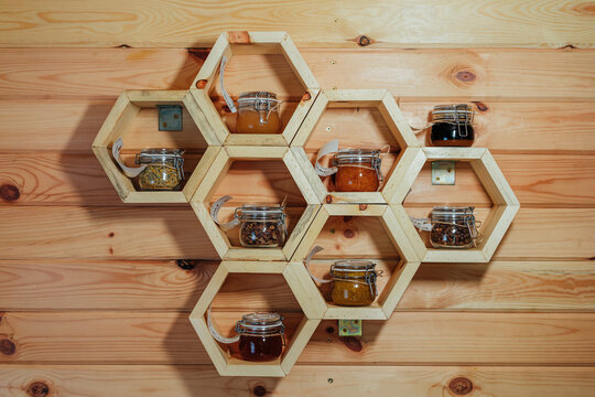 Glass jars containing assorted honey, bee pollen, and other bee products displayed on hexagonal honeycomb-shaped wooden shelves against a rustic timber wall