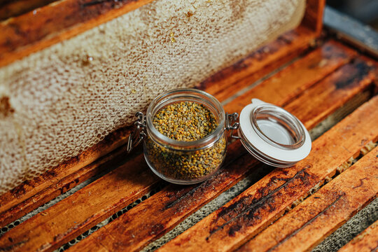 Close-up of colorful bee pollen granules stored in an open glass jar beside honeycomb frames on a rustic wooden beehive