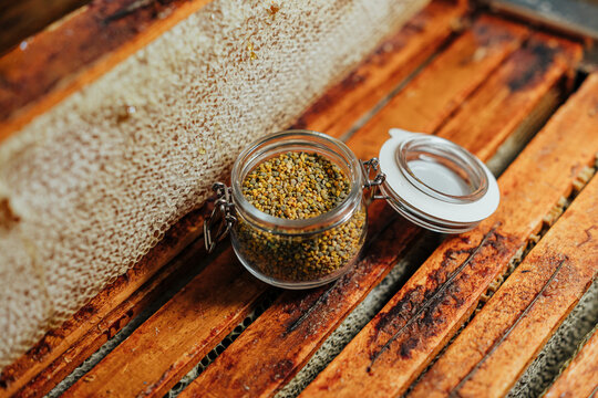 Close-up of colorful bee pollen granules stored in an open glass jar beside honeycomb frames on a rustic wooden beehive