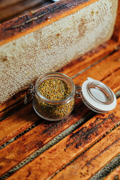 Close-up of colorful bee pollen granules stored in an open glass jar beside honeycomb frames on a rustic wooden beehive