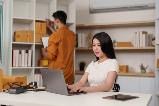 Asian woman working on laptop in ecommerce office with colleague checking inventory