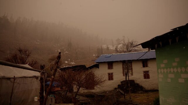 A two story stone house with a blue metal roof stands beside a green painted building as fog drifts over a snow patched hillside