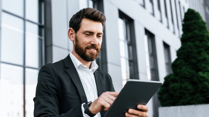 Fototapeta premium Focused Caucasian Man In Business Suit Using Tablet While Standing Outside Modern Urban Office Building, Engaged In Work. Professional Setting Emphasizing Business Concept.
