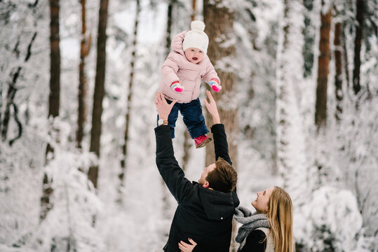 Mom and dad smiling, enjoying winter walk in nature. Baby's laughter and joy. Happy parents playing with daughter, tossing her high up in snowy forest. Close up.