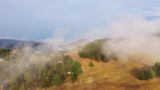 Aerial view of La Molina ski resort in the Spanish Pyrenees, with low clouds drifting over forested mountain slopes, ski lifts and alpine village visible below