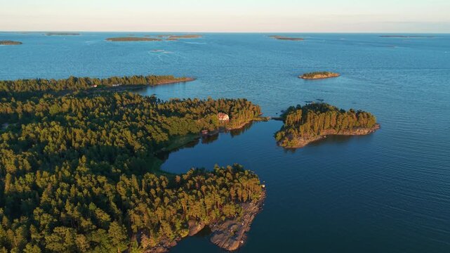 Aerial view of Helsinki archipelago at golden hour, featuring numerous forested islands, a lakeside cottage, calm blue Baltic Sea waters, and scattered islets stretching to the horizon in Finland