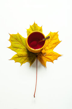 Minimalist flat lay autumn background - a yellow ceramic mug with tea sitting on a single autumn maple leaf over a white background