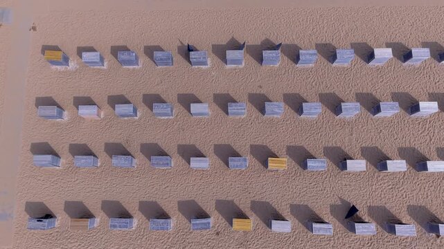 Aerial view video clip of Katwijk aan Zee beach resort top down, wooden beach cabins casting shadows, geometric grid pattern, and golden sand in South Holland, Netherlands