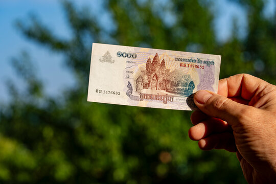 Hand holding Cambodian riel banknote outdoors, closeup. Person's hand holding 1000 Cambodian riel paper currency note featuring Angkor Wat temple design against blurry green nature background