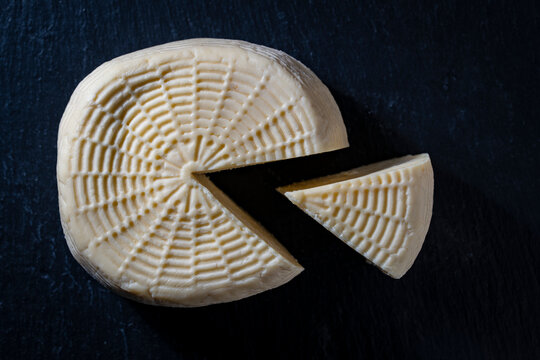 Homemade bryndza cheese head with cut slice, closeup. Fresh soft round cheese or sulguni head with one slice cut out showing wicker mold texture on dark slate background shot from above