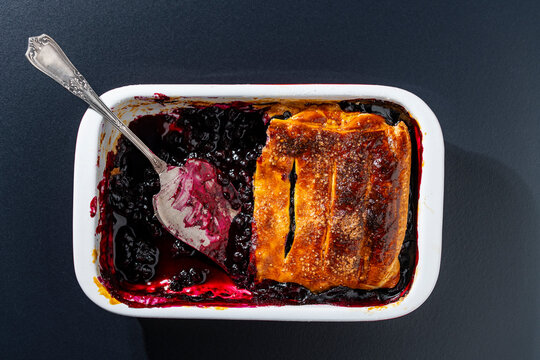 Homemade blueberry pie in baking dish, top view. Freshly baked blueberry cobbler pie with golden crispy crust in white ceramic pan with vintage metal serving spatula on dark background, closeup