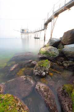 View of the Costa dei Trabocchi with a traditional wooden fishing structure extending over the calm sea, featuring moss-covered rocks in the foreground in Marina di San Vito, Abruzzo, Italy.