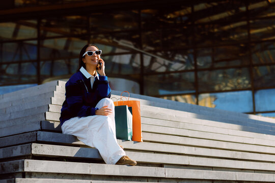 Happy woman sitting on concrete stairs chatting on her smartphone with shopping bags beside her on a sunny urban day