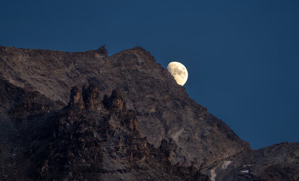 View of the moon rising behind the rocky Chastelets Peaks under a deep blue twilight sky in Silvaplana, Grisons, Switzerland.