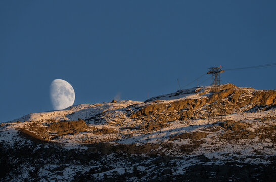 View of the moon rising over the snow-covered Chastelets Peaks with a cable car pylon silhouetted against the clear blue sky during golden hour in Silvaplana, Grisons, Switzerland.