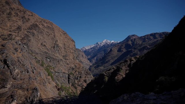 Dramatic rocky canyon with steep sunlit and shadowed cliffs framing a distant snow capped Himalayan peak under a deep clear blue sky, Api Himal Farwest Nepal