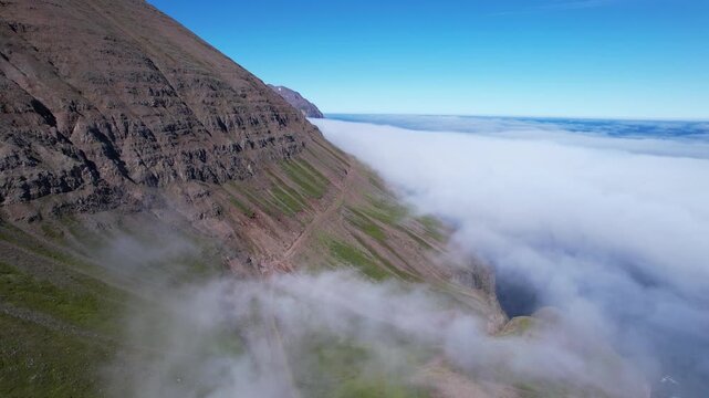 Aerial drone view of steep volcanic mountain slopes rising above a cloud filled valley in Iceland featuring rugged ridgelines drifting clouds and clear blue sky