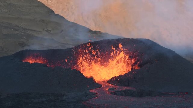 Slow motion aerial drone view of an active volcanic eruption in Iceland showing bright molten lava surging from a volcanic vent flowing across dark basalt with rising steam