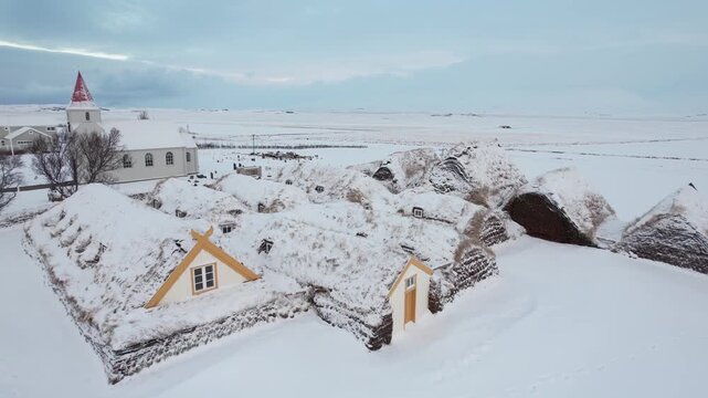 Aerial drone view of snow covered traditional turf houses and a church in rural Iceland featuring winter landscape open fields historic architecture and blue sky