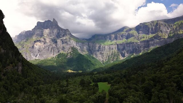 Mountain cliffs tower over the forested Cirque du Fer a Cheval in France, with thin waterfalls descending rock walls and low clouds covering the upper ridges in broken sunlight.