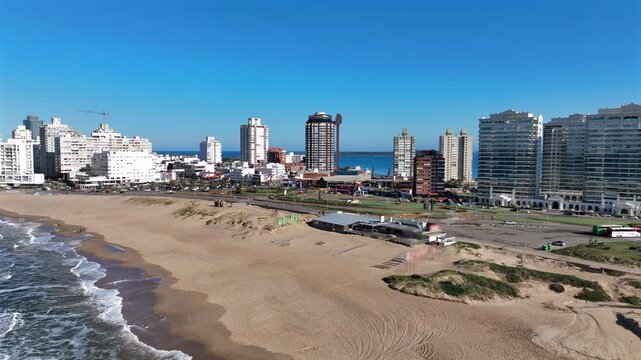 Playa Brava beach truck right along the Punta Del Este waterfront, passing the iconic Los Dedos hand sculpture with city skyline and sand dunes under sunny sky.