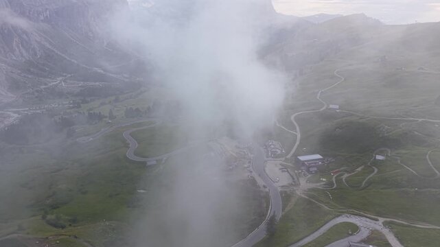 Cinematic drone view above Gardena Pass as drifting clouds partially conceal winding alpine roads and rugged mountain scenery in the Dolomites, Italy