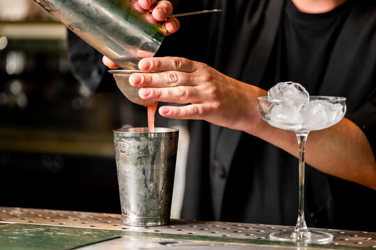 A bartender straining a cocktail from a metal shaker tin into a metal glass through a fine mesh strainer next to a coupe glass filled with ice on a bar counter.