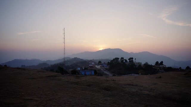 Silhouette of a tall telecom tower on a hilltop overlooking a golden sunset behind layered Himalayan foothills and a small mountain village